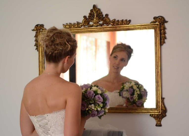 Sposa in abito bianco davanti a uno specchio dorato, bouquet floreale e acconciatura elegante