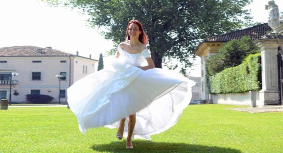 Sposa in abito bianco che corre in un giardino soleggiato, circondata da siepi e edifici rustici.