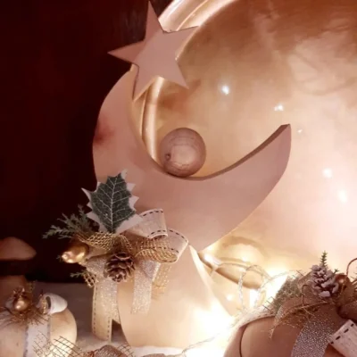 Dettaglio di angioletto o decorazione a mezzaluna in legno per il Natale, ornato con nastro dorato, pigna e agrifoglio artificiale, su sfondo illuminato.
