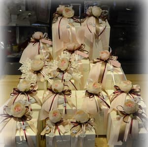 Confezioni regalo artigianali con fiocchi rosa e bordeaux, rose decorative e dettagli in pizzo bianco, disposte su alzate eleganti.
