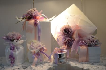 Bomboniere artigianali con fiocchi pastello, fiori decorativi rosa e lilla, perle e nastri su superficie in pizzo, ideali per eventi eleganti.