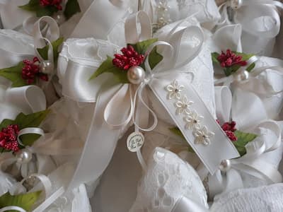 Sacchettino bomboniera artigianale con nastri bianchi e rosa, bacche rosse, foglie verdi e perle decorative su tessuto bianco.