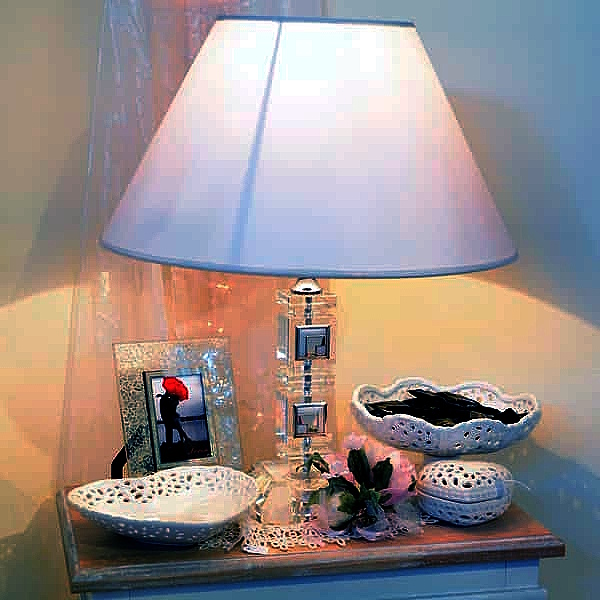 Lampada con base trasparente e paralume bianco su tavolo decorato con ciotole traforate, fiori artificiali e fotografia incorniciata su raffinata cornice.