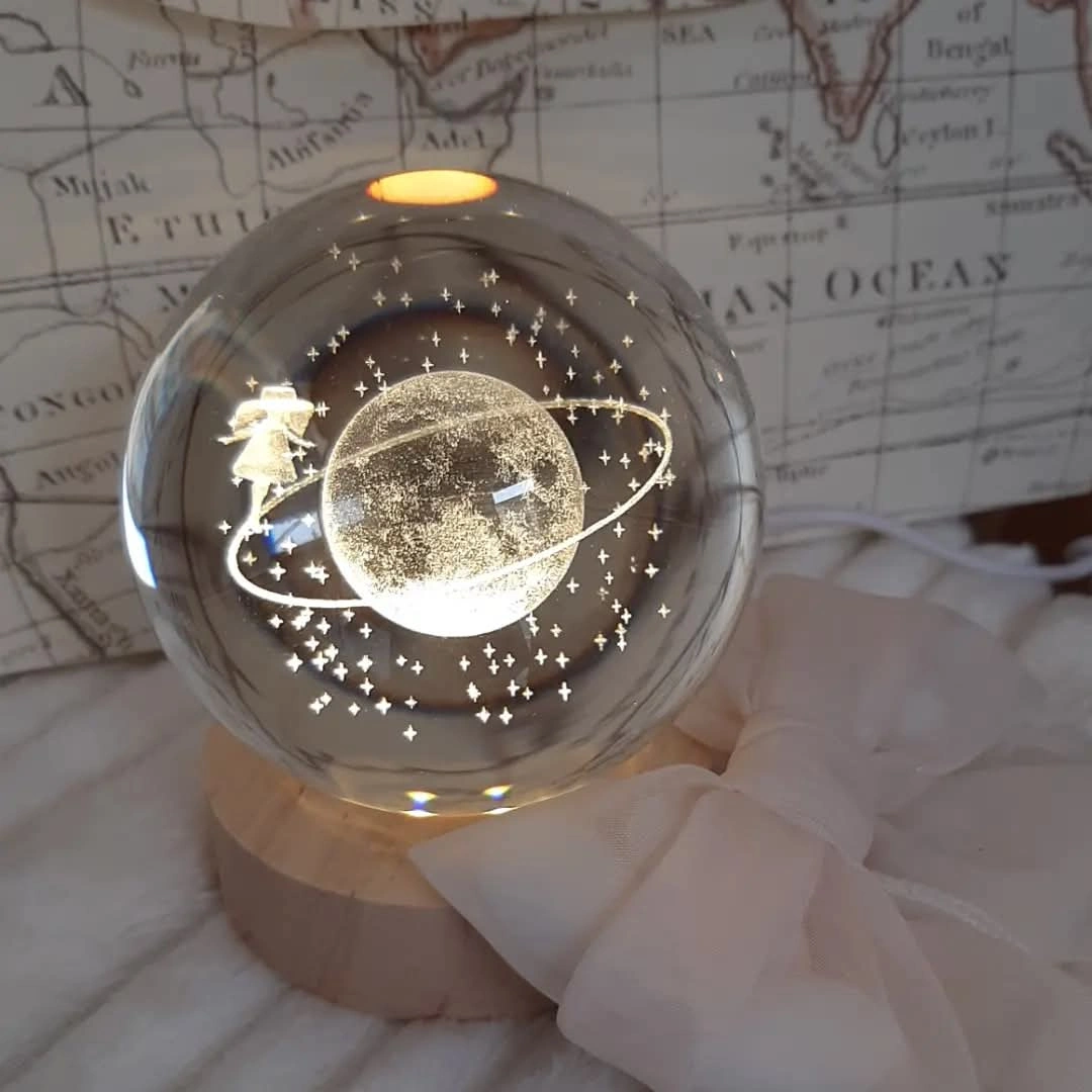 Globo di vetro trasparente con incisione 3D laser del Piccolo Principe su un pianeta con anello e stelle, poggiato su base LED in legno.